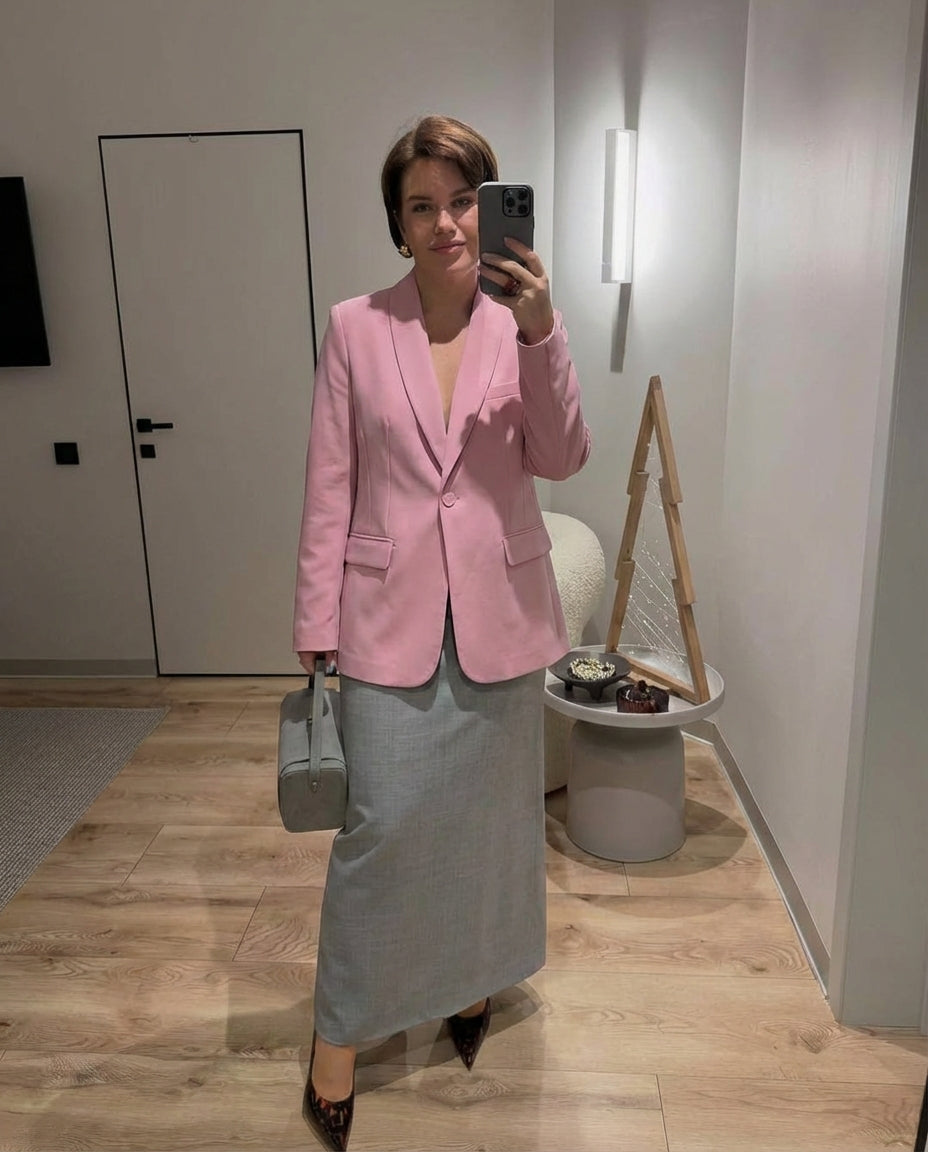Soft pink tailored blazer with a shawl lapel and single-button closure, styled with a grey maxi skirt and handbag.