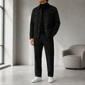 Man wearing a black casual button-front jacket with matching black trousers, a black turtleneck sweater, and white sneakers in a modern indoor setting.
