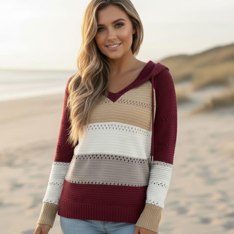 Color-block striped knit hooded pullover with eyelet crochet trim and a relaxed V-neck, worn on a sandy beach.