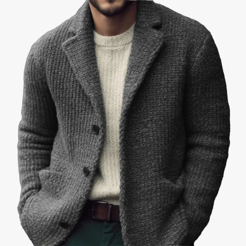 Charcoal grey textured knit cardigan with a notch collar, button-front closure, and patch pockets over a cream sweater.