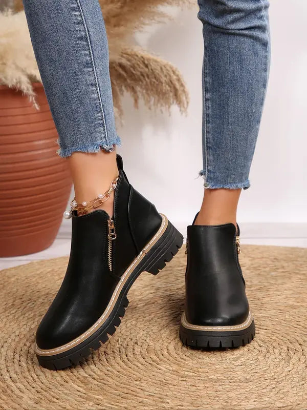 Black brushed-finish ankle boots with side zips, round toes, and chunky notched tread soles worn with blue denim jeans.
