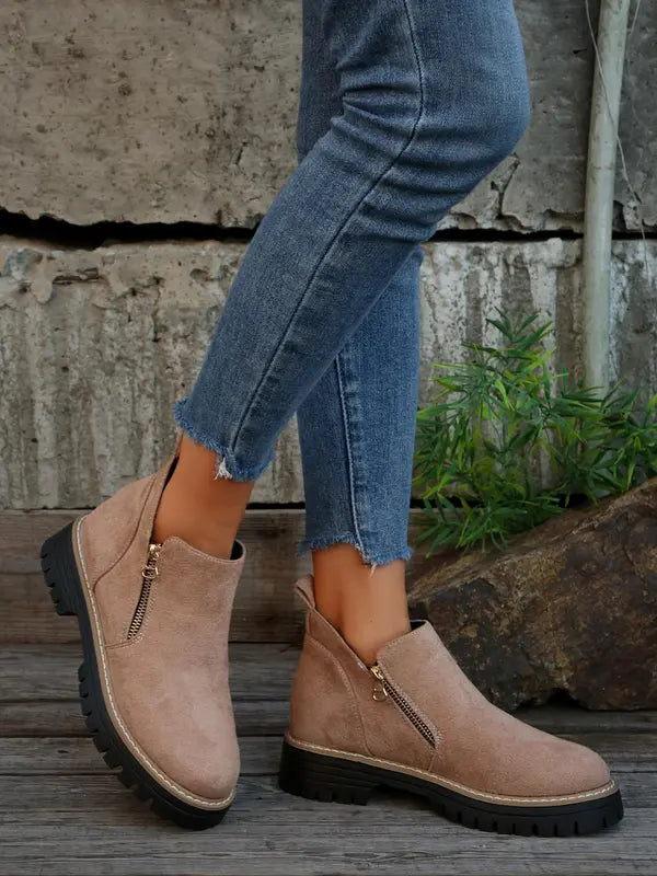 Taupe brushed-finish ankle boots with side zips, round toes, and thick notched tread soles worn with cropped blue jeans.