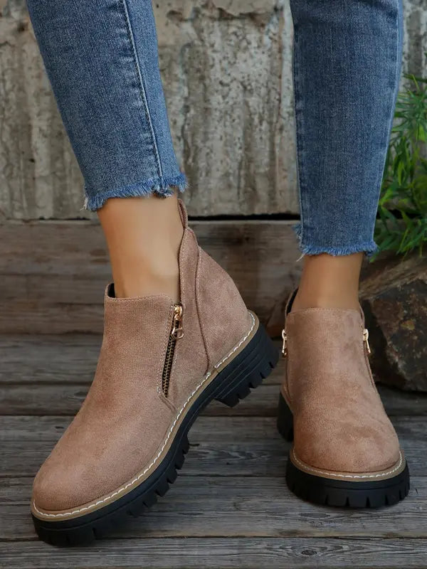 Taupe brushed ankle boots with side zips, round toes, and thick black notched tread soles worn with frayed blue jeans.