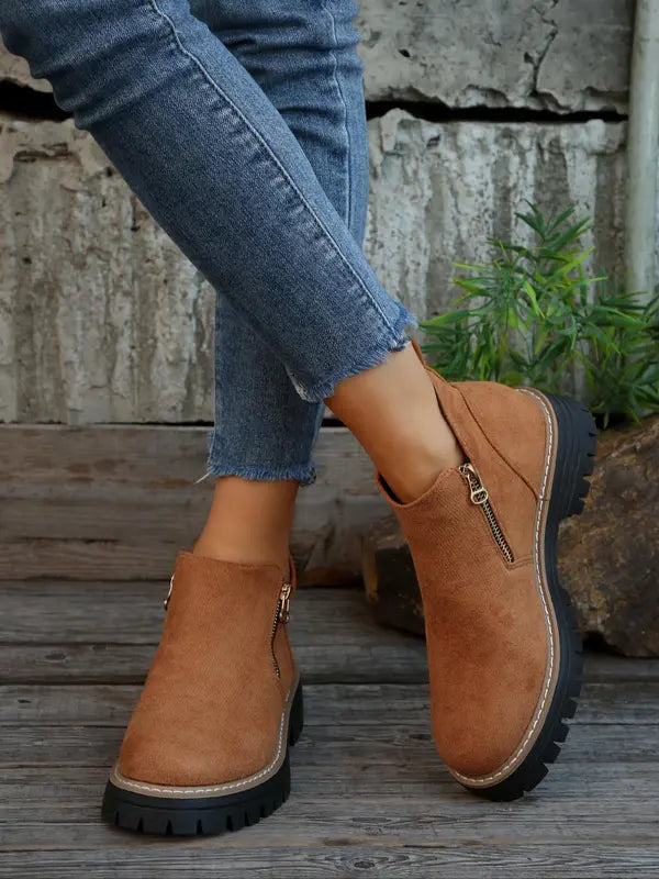 Tan brushed ankle boots with side zips, round toes, and thick black notched tread soles worn with cropped blue jeans.