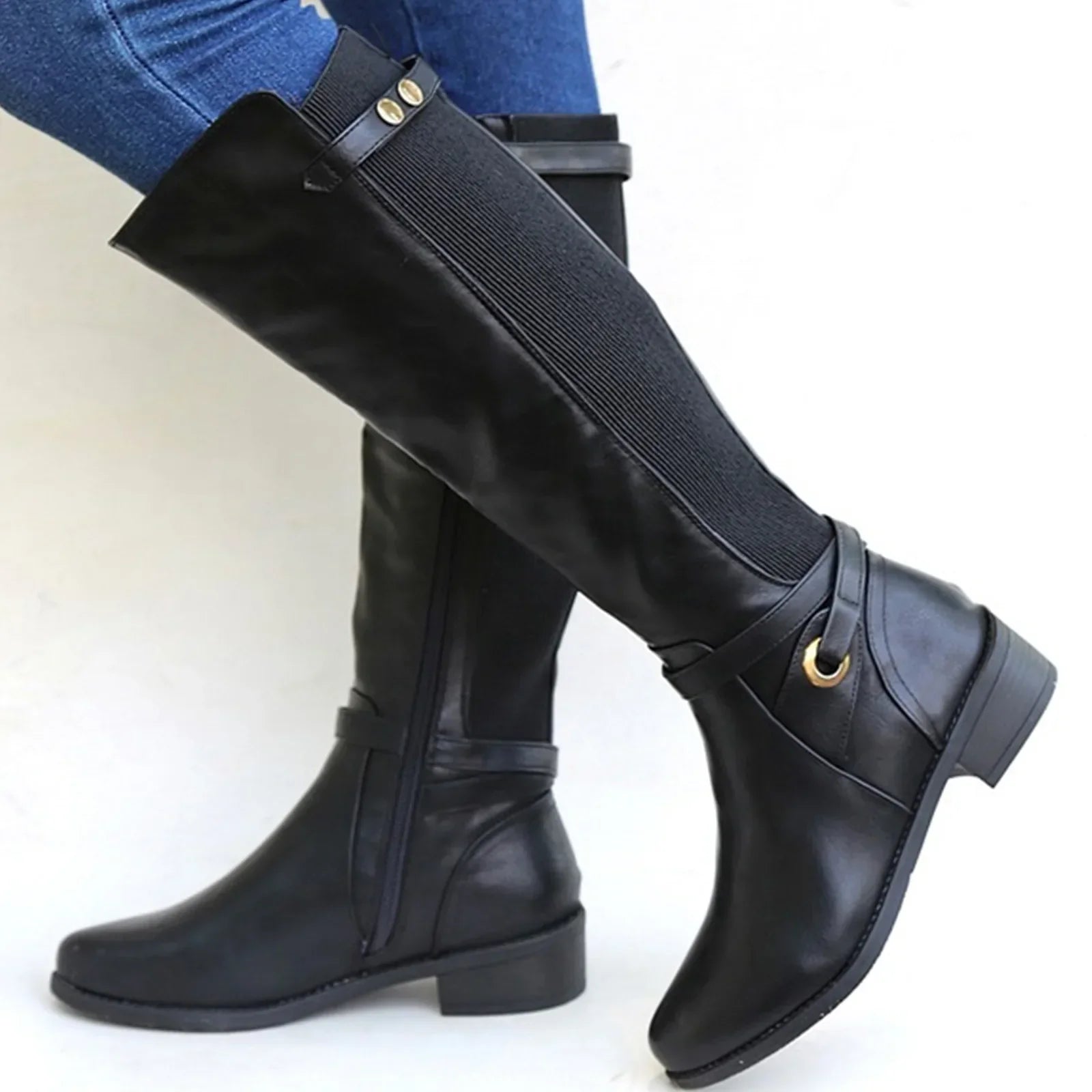 Black knee-high leather riding boots featuring a side zipper, elastic back panel, decorative buckle strap, and low block heel, styled with blue jeans.