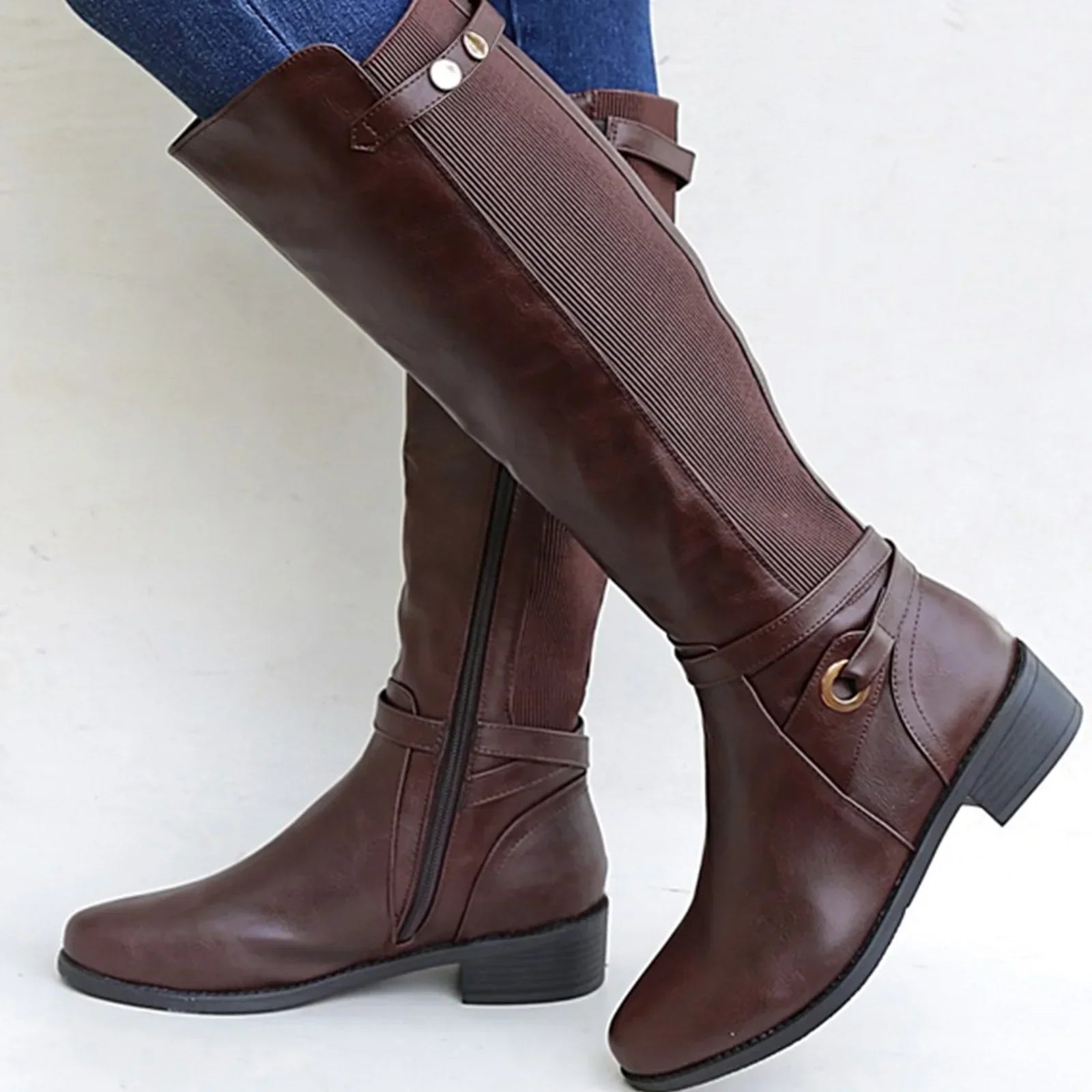 Brown knee-high leather riding boots with a side zipper, elastic back panel, decorative buckle strap, and low block heel, worn with blue jeans.