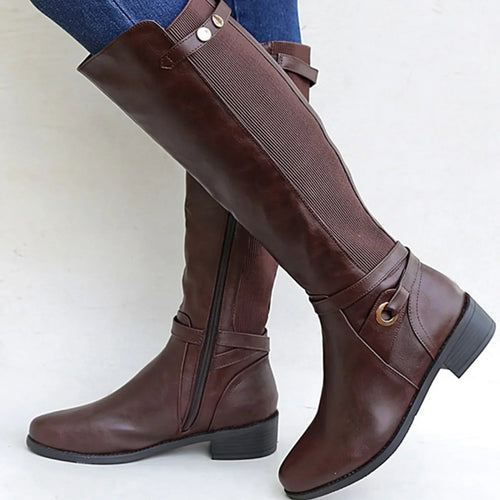 Brown knee-high leather riding boots with a side zipper, elastic back panel, decorative buckle strap, and low block heel, worn with blue jeans.