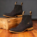 Black suede western ankle boots with pointed toes, elastic side panels, and stacked wooden heels on a rustic wood block.