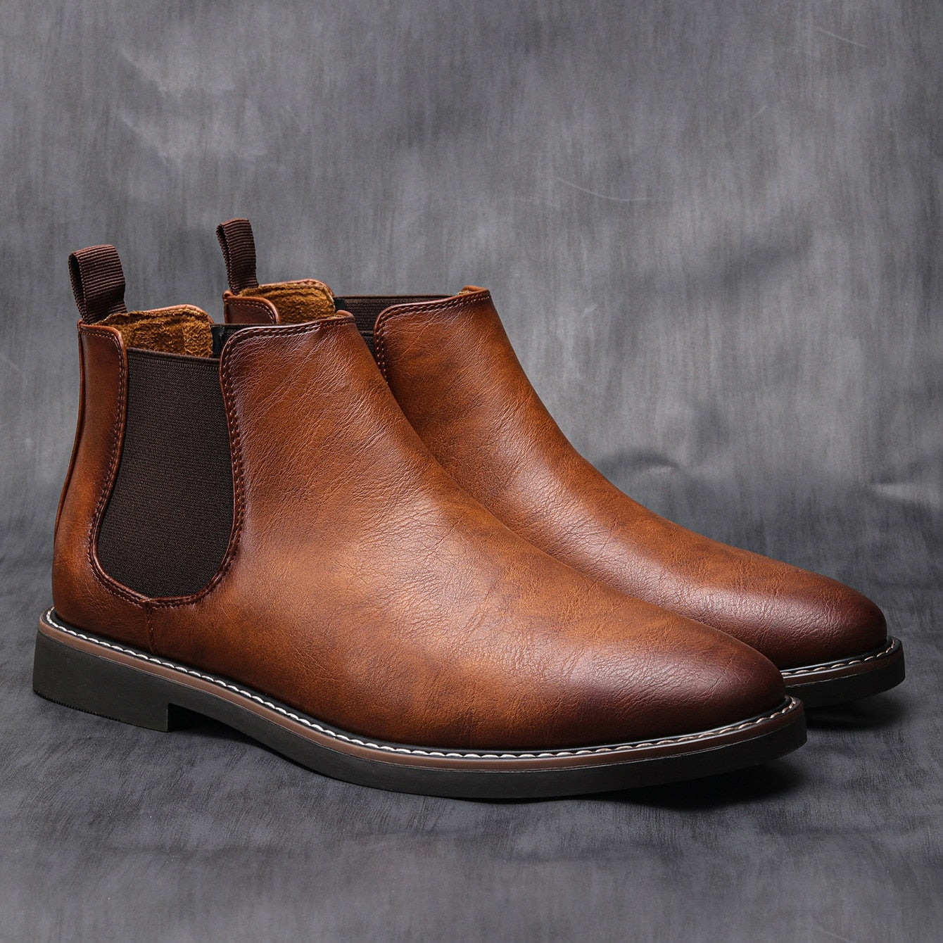 Tan leather Chelsea boots with a round toe, elastic side gores, pull tabs, and a stitched welt on a dark grey background.