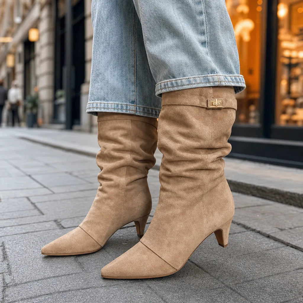 Beige slouch knee-high suede boots with pointed toes and mid block heels, paired with light blue jeans on a city sidewalk.