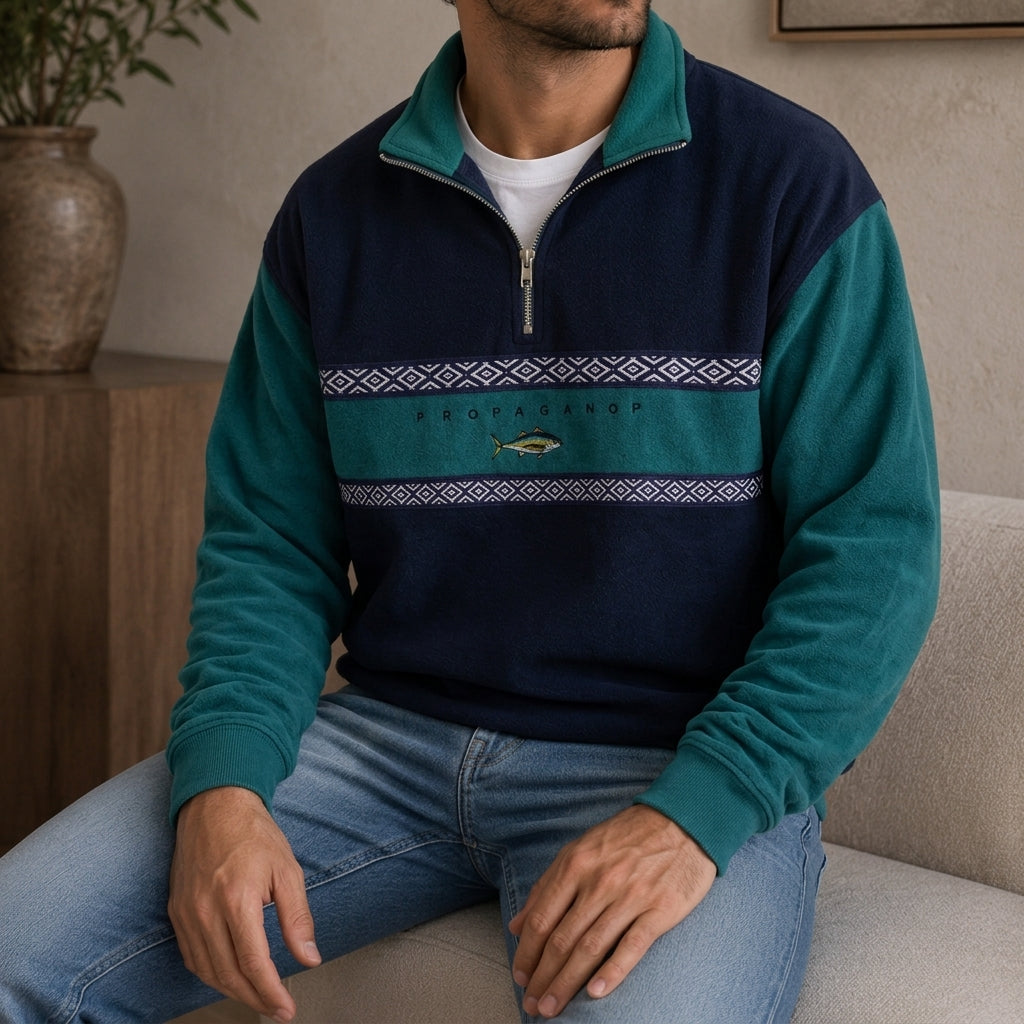 Teal and navy colorblock quarter-zip pullover worn by a male model, featuring a geometric chest band and embroidered motif.