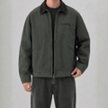 Front view of a model wearing a dark washed-green cotton canvas barn jacket with a dark brown corduroy collar, zippered chest pocket, welt hand pockets, and full zip closure.