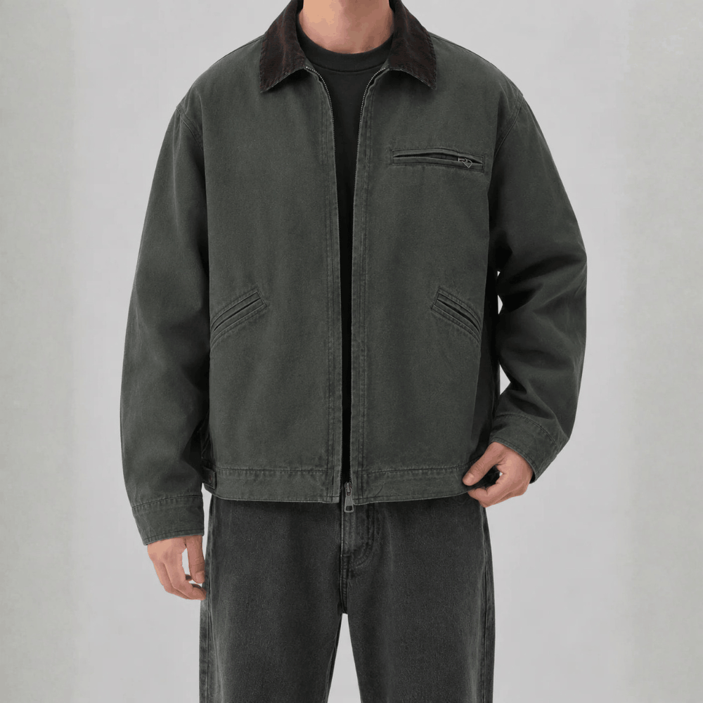 Front view of a model wearing a dark washed-green cotton canvas barn jacket with a dark brown corduroy collar, zippered chest pocket, welt hand pockets, and full zip closure.