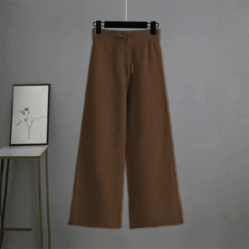 Front view of the high-waisted dark brown wide-leg knit trousers hanging against a grey wall background.