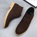 Dark brown suede desert boots with a plain toe, lace-up closure, and a flat, light-colored wedge sole with welt stitching.
