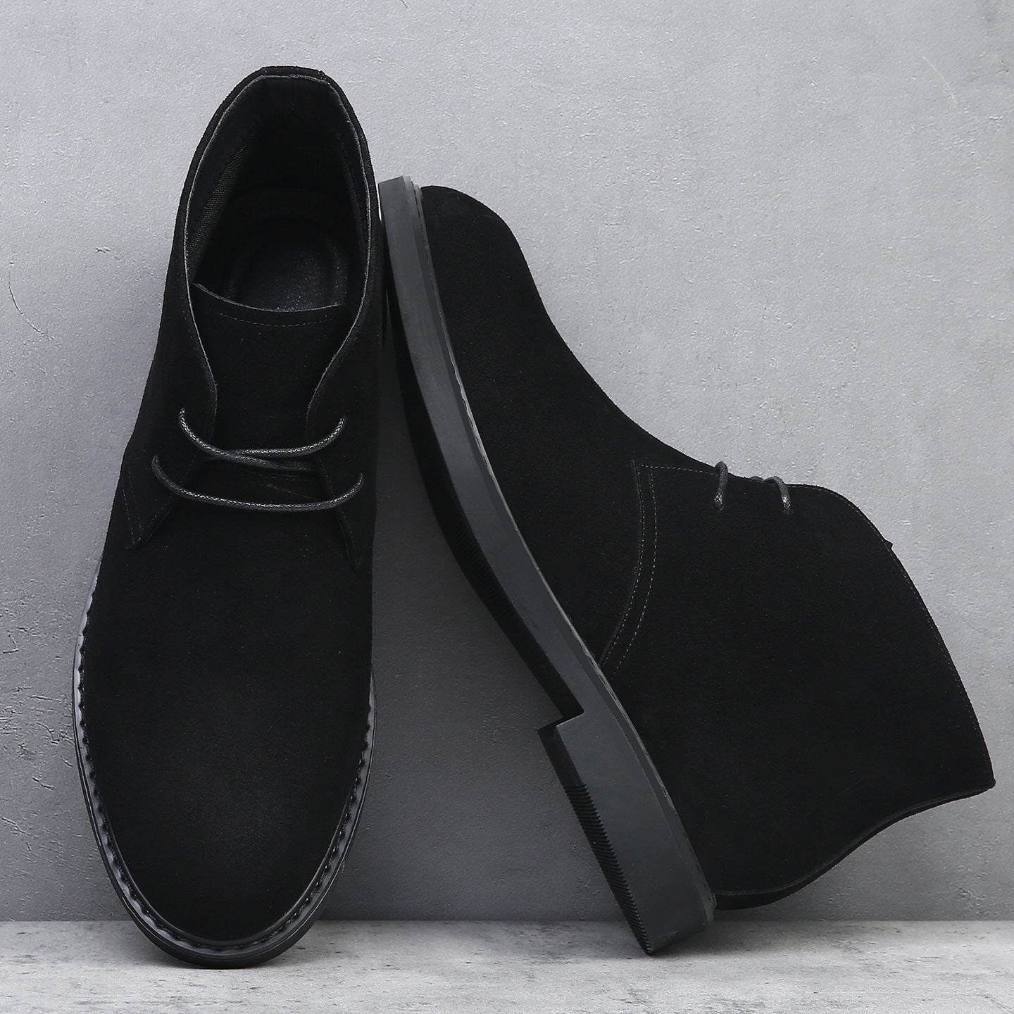 Black suede desert boots featuring a plain toe, lace-up closure, and a flat, tonal wedge sole with visible welt stitching.
