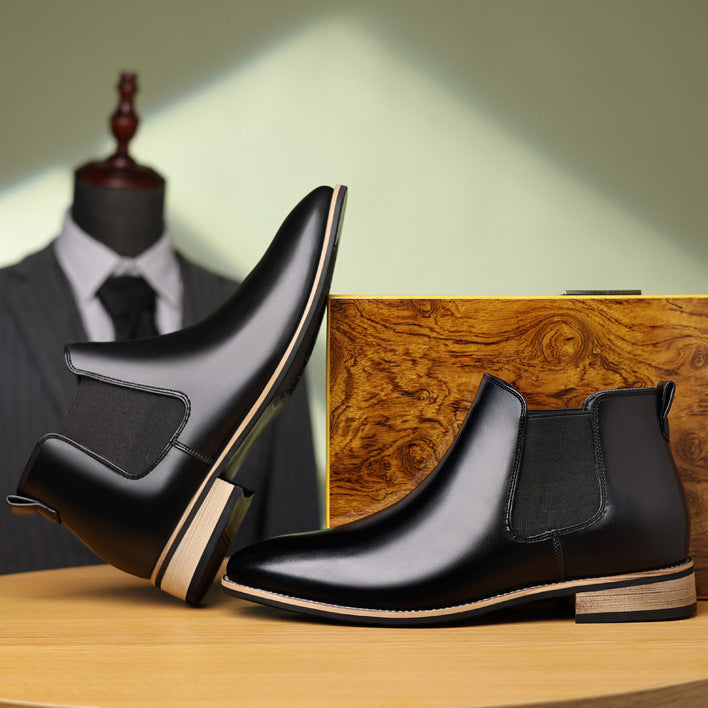 Black leather Chelsea boots with pointed toes, elastic side panels, and light-toned stacked heels on a wooden surface.