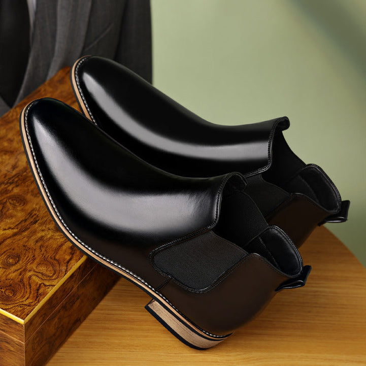 Burnished black leather Chelsea boots with a pointed toe, stitched welt, and low stacked heel resting on a wooden surface.