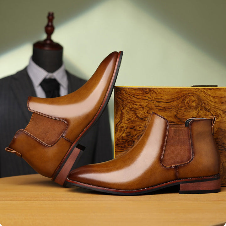 Burnished tan leather Chelsea boots with pointed toes, elastic side panels, and low stacked heels on a wooden surface.