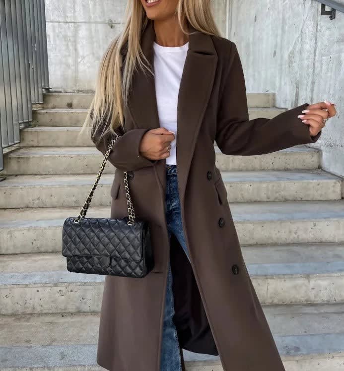 Dark brown double-breasted longline coat with notched lapels and welt pockets, worn over a white top and blue jeans.