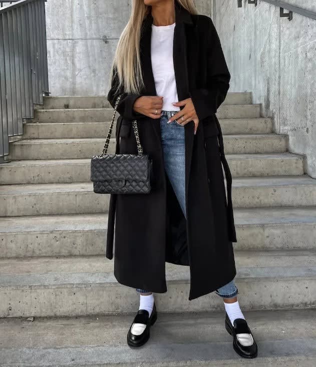 Black longline coat with notched lapels and a smooth matte finish, worn open over a white tee, jeans, and loafers.