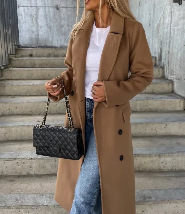 Camel double-breasted longline coat with notched lapels and welt pockets, worn over a white tee and blue jeans.
