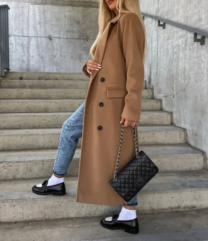 Camel double-breasted longline coat with notched lapels and welt pockets, worn over blue jeans and black loafers.