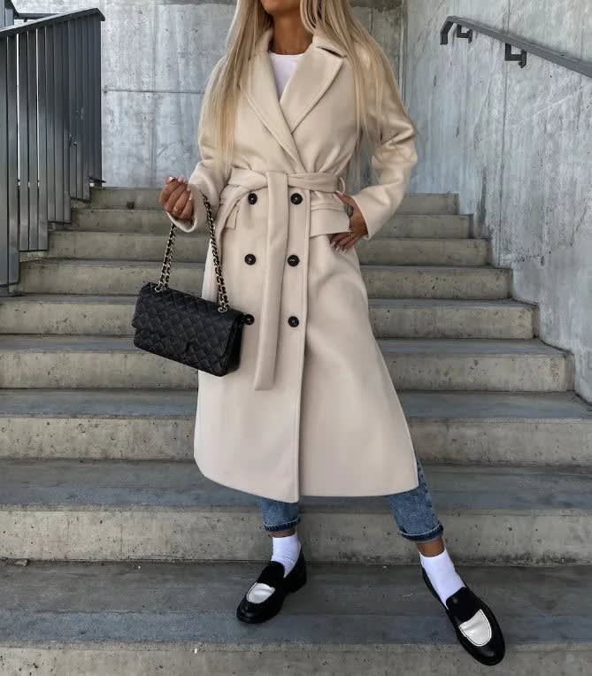 Beige double-breasted longline coat with notched lapels, tie waist, and welt pockets, worn over jeans and loafers.
