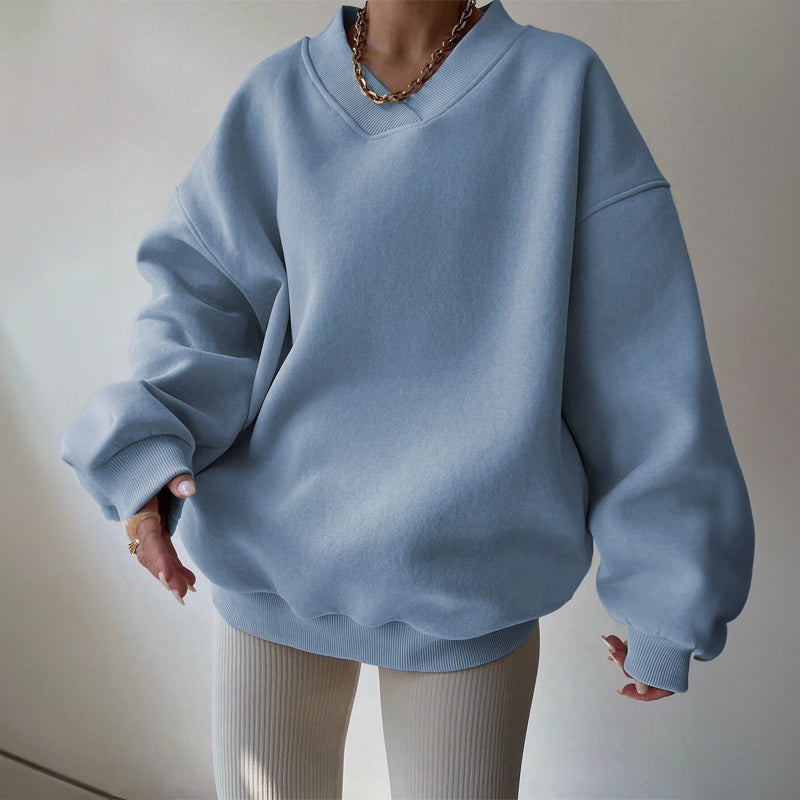 Blue oversized V-neck sweatshirt with dropped shoulders, ribbed cuffs, and a relaxed fit, styled with light-colored pants.