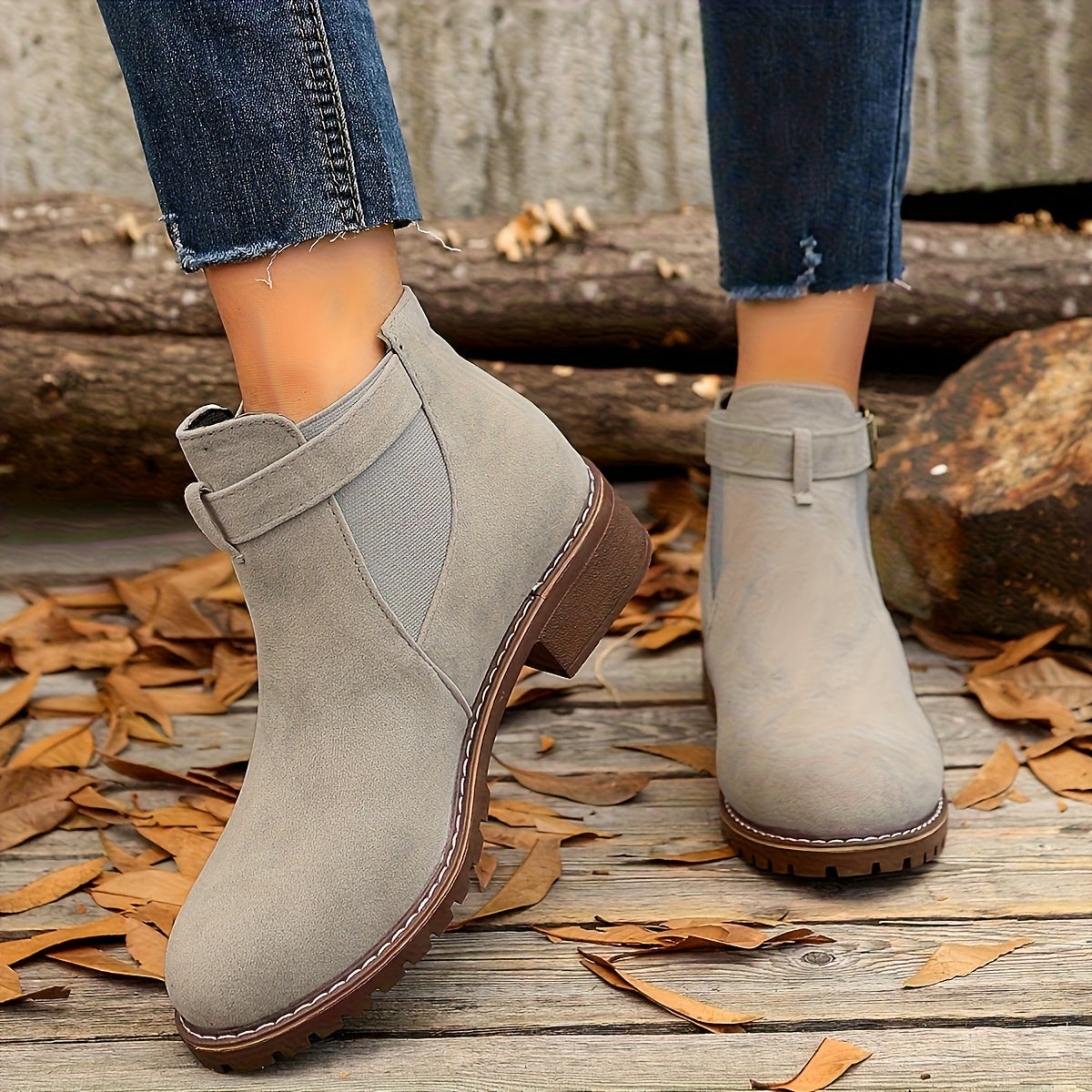 Grey suede Chelsea boots with buckle strap, elastic side panels, and a rugged brown lug sole on a wooden surface.