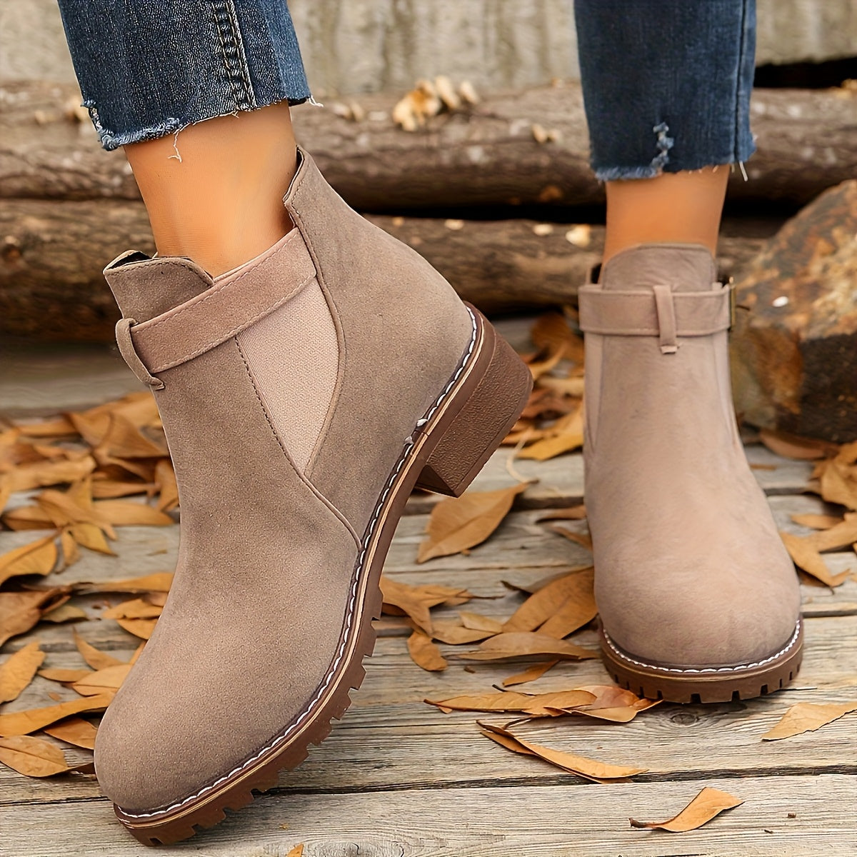 Taupe suede Chelsea boots with a buckled ankle strap, elastic side panels, and a rugged lug sole on a wooden surface.