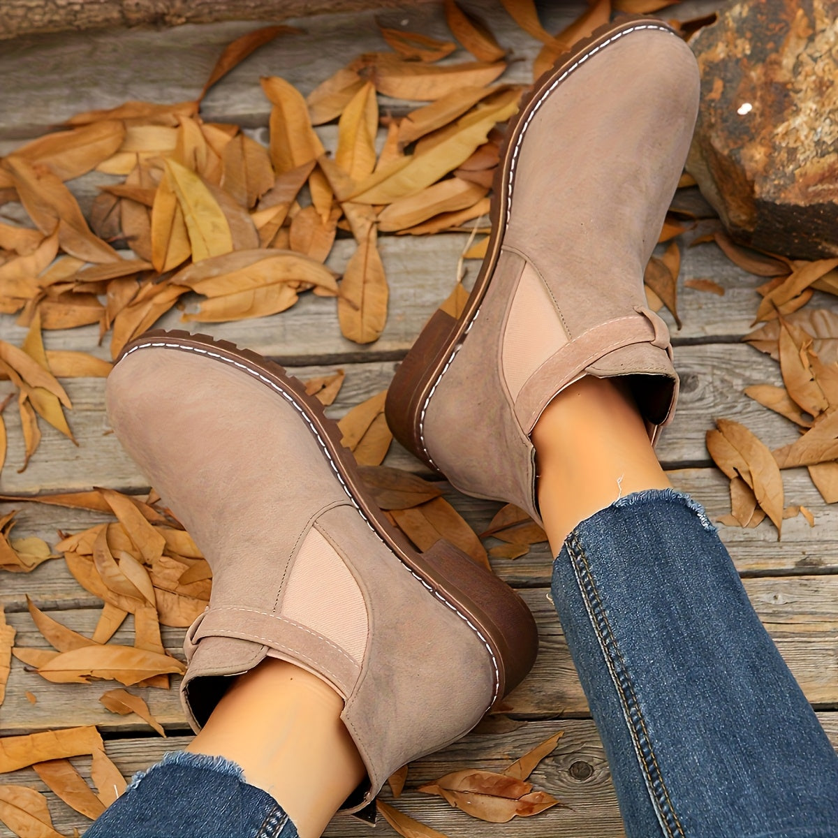Taupe suede Chelsea boots with elastic side panels, buckle strap detail, and a rugged lug sole on a wooden surface.