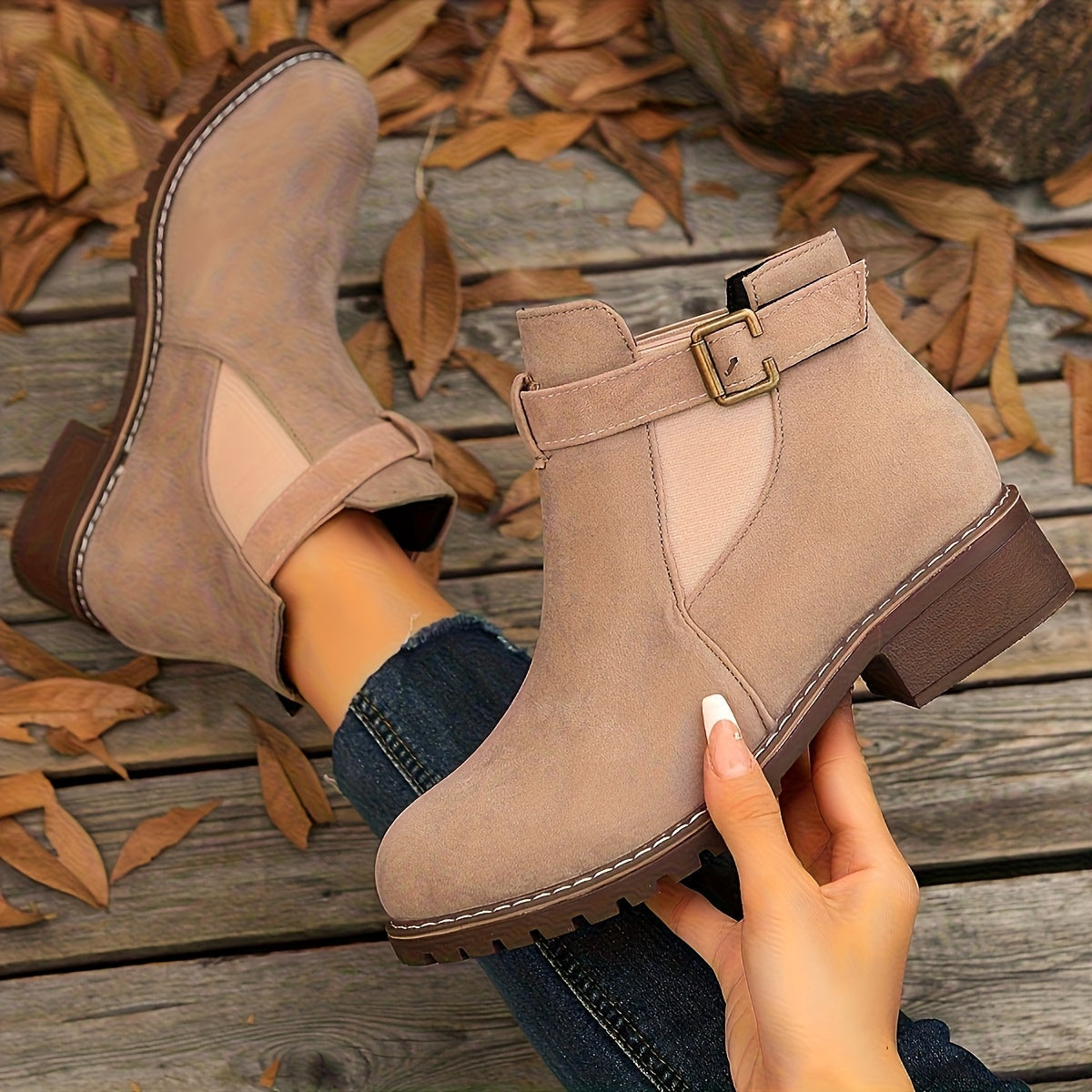 Taupe suede Chelsea boots with a buckle strap, elastic side panels, and a rugged lug sole block heel on a wooden surface.