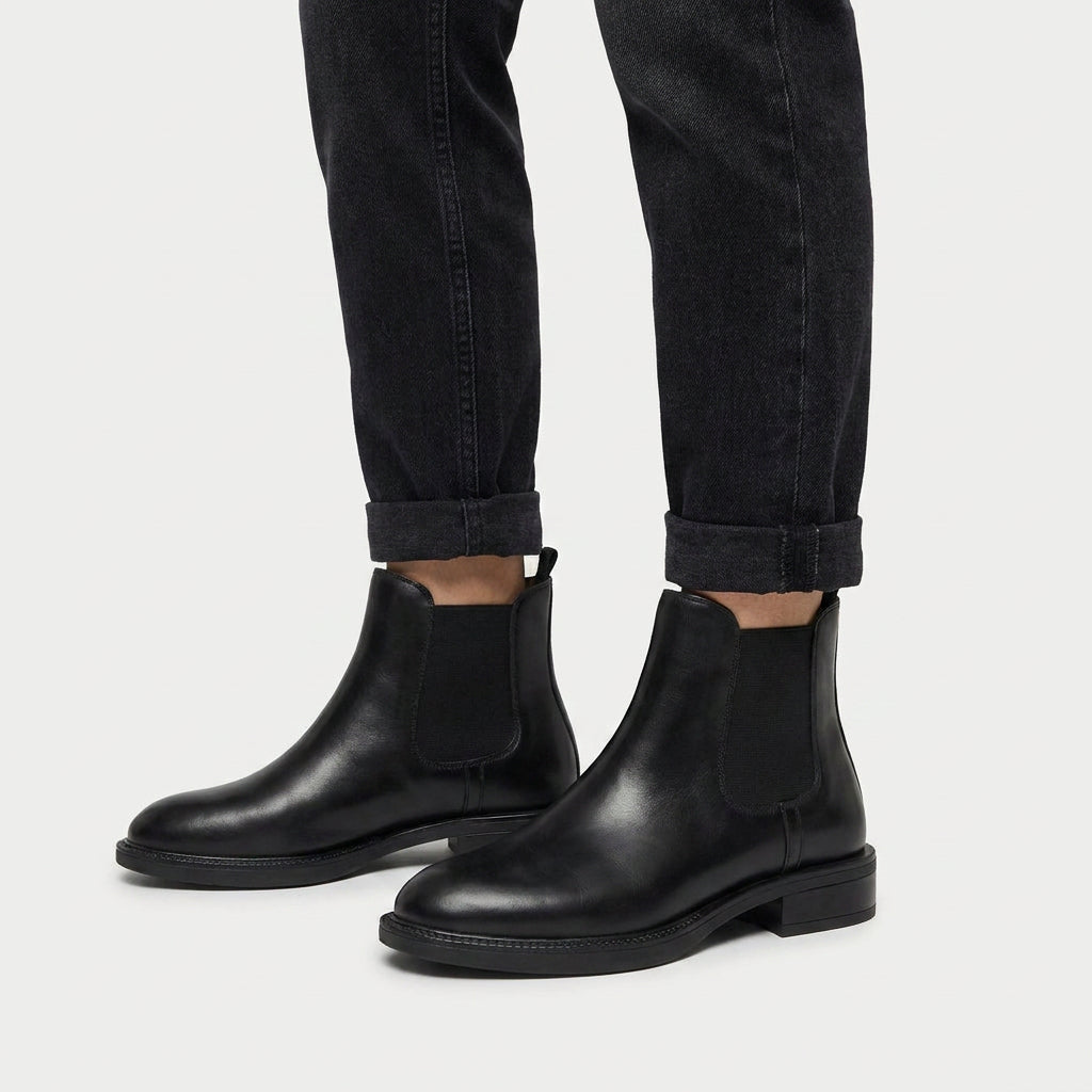 Black vegan leather Chelsea boots with elastic side panels, a rear pull tab, and a low stacked heel worn with cuffed jeans.