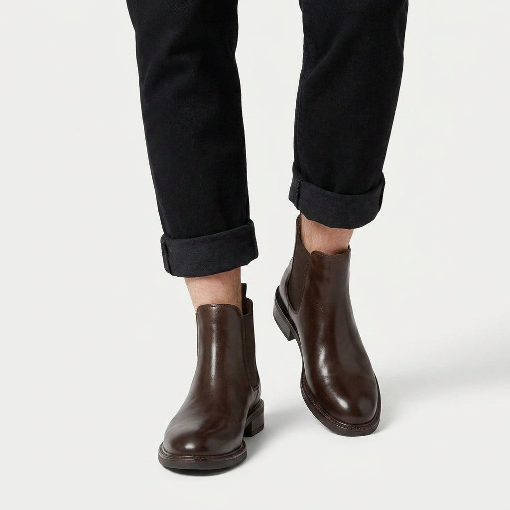 Dark brown vegan leather Chelsea boots with elastic side panels and a low stacked heel worn with black cuffed trousers.