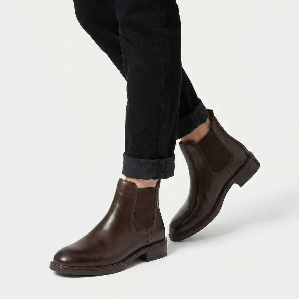 Dark brown vegan leather Chelsea boots with elastic side panels and a low stacked heel worn with cuffed black trousers.