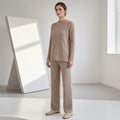 Full-length front view of a model in a minimal studio setting wearing a taupe beige ribbed knit lounge set featuring a mock-neck sweater with side slits and matching wide-leg pants, styled with flat loafers.