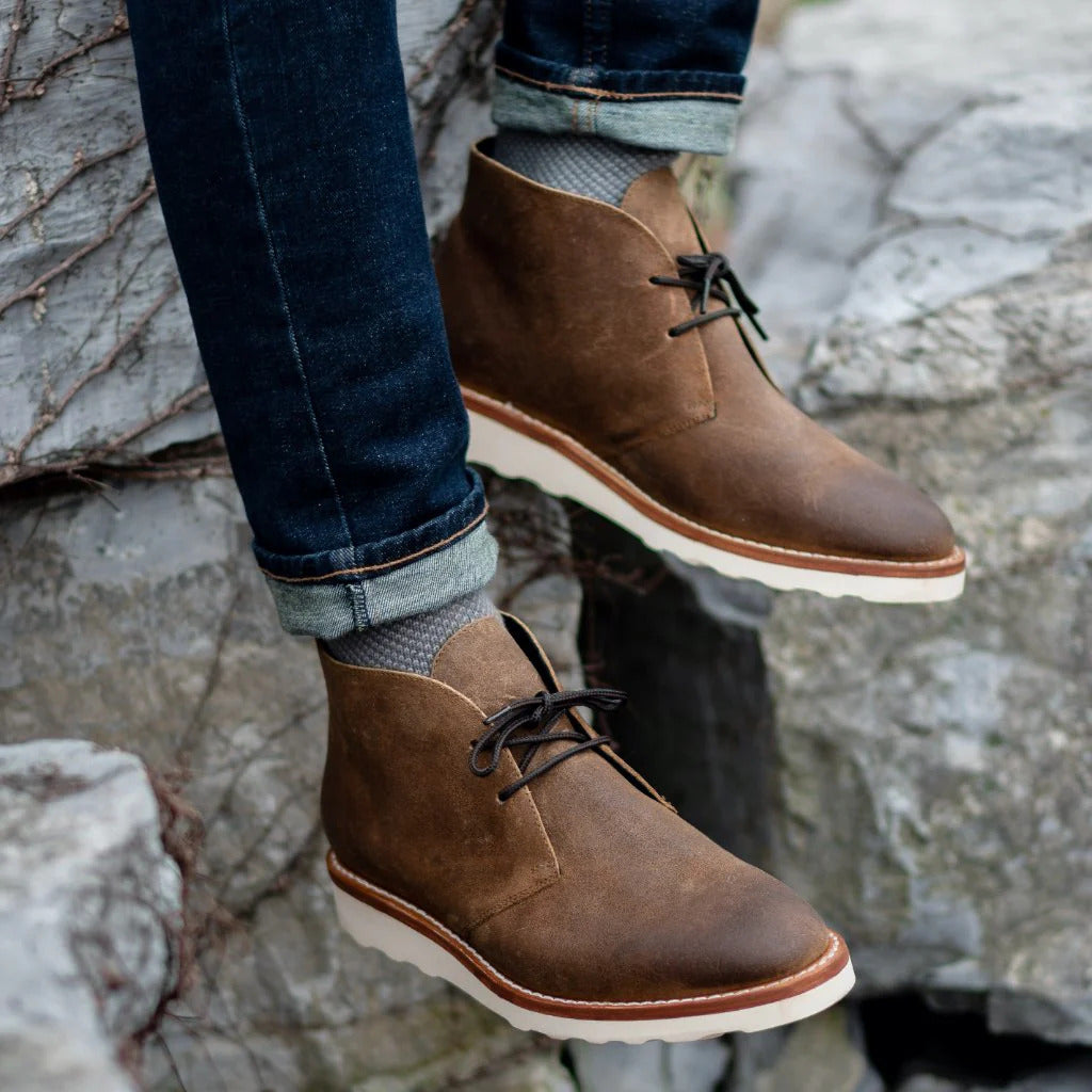 Brown napped leather chukka boots with two-eyelet lacing, white treaded outsoles, and visible welt stitching worn with denim.