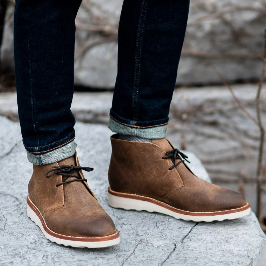 Brown napped leather chukka boots with two-eyelet lacing, white treaded outsoles, and visible welt stitching worn with denim.