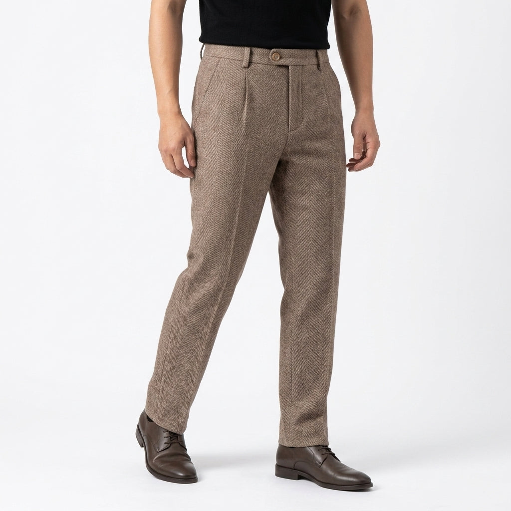 Front three-quarter view of a man wearing beige herringbone wool trousers with a pleated front, tailored fit, slant pockets, and brown dress shoes, paired with a black t-shirt against a white background.