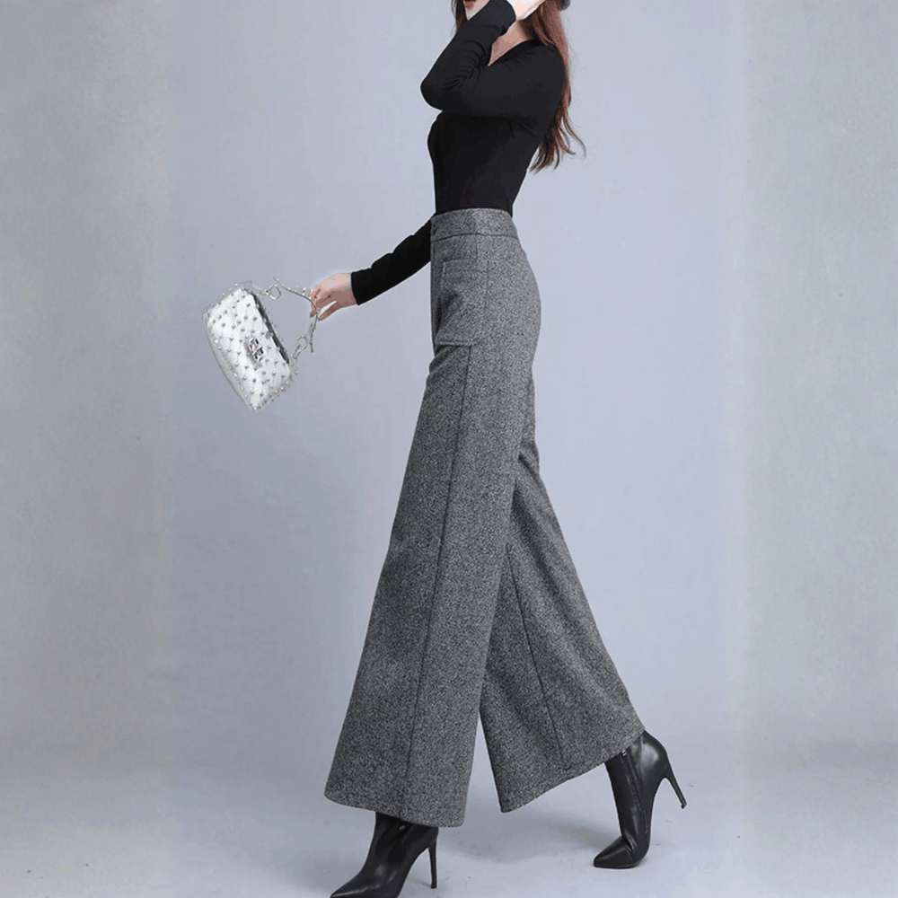 Side profile view of a model in motion wearing high-waisted gray wide-leg trousers paired with black heeled boots.