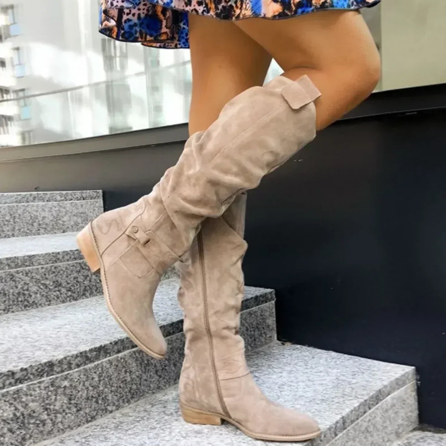 Beige slouch knee-high suede boots featuring pointed toes and block heels, worn while walking up outdoor steps.