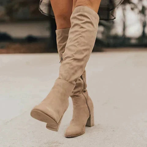 Beige slouch knee-high suede boots featuring a relaxed shaft, pointed toes, and block heels, worn outdoors.