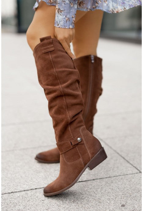 Brown slouch knee-high suede boots featuring pointed toes, side buckle accents, and block heels, worn with a floral dress.