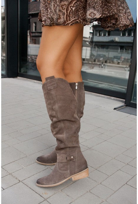 Gray slouch knee-high suede boots featuring pointed toes, side buckle accents, and block heels, worn outdoors.