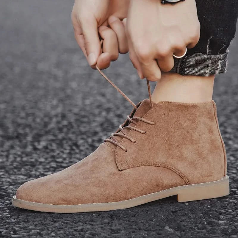 Tan suede chukka boots with a plain toe, lace-up front, and welted sole edge being tied on dark denim.