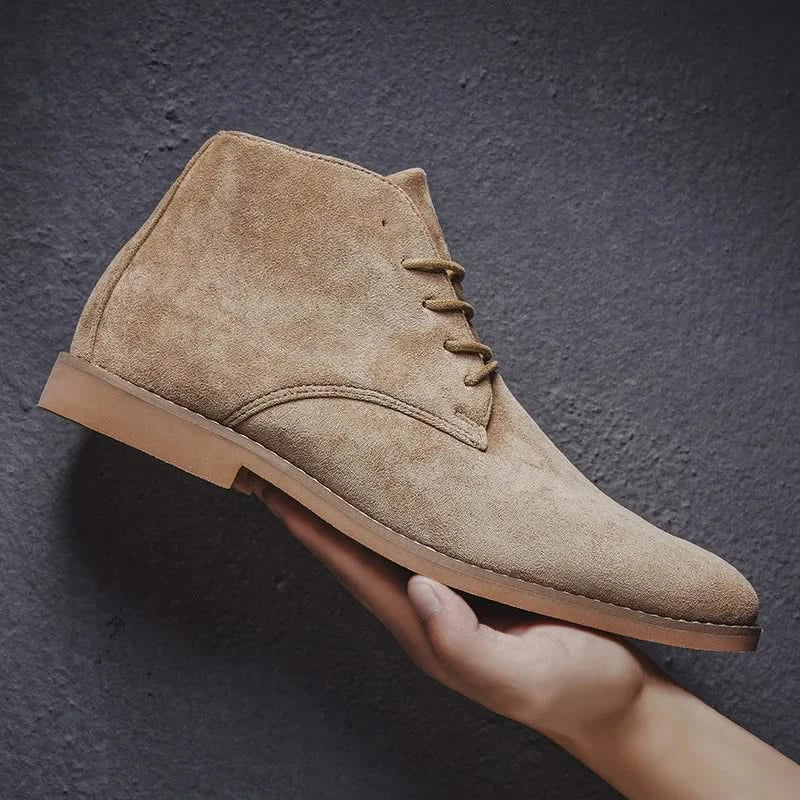 Tan suede chukka boot with a plain toe, lace-up front, and low block heel held against a dark textured background.