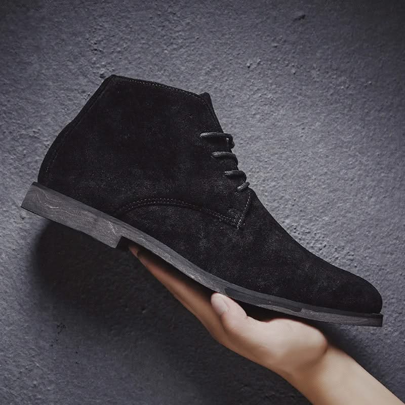 Black suede chukka boot with a plain toe, lace-up front, and a low block heel held against a textured dark background.