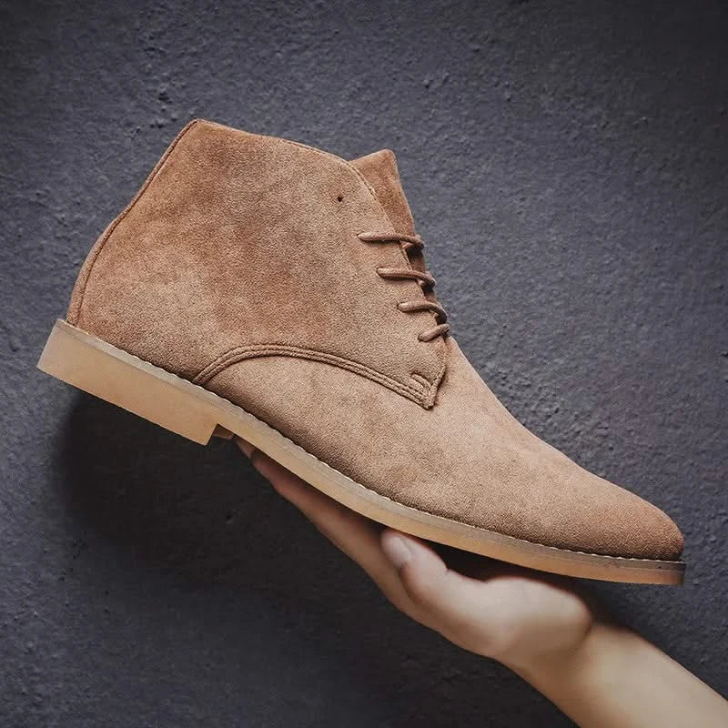 Tan suede chukka boots with a plain toe, lace-up front, and low block heel held against a dark textured background.
