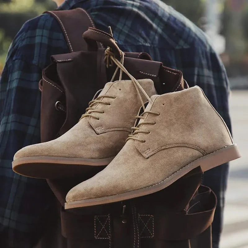 Tan suede chukka boots with a plain toe, lace-up front, and welted sole edge hanging from a leather backpack.